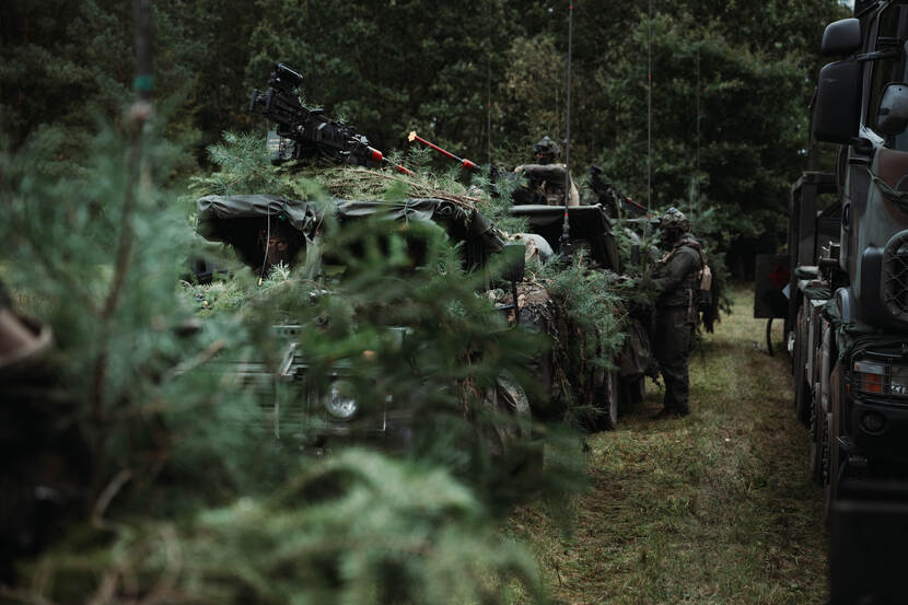 Op een vrachtwagen staan houten kisten met daarin gevechtrantsoenen en water. Twee militairen bevoorraden een voertuig met daarin twee gemaskerde militairen.