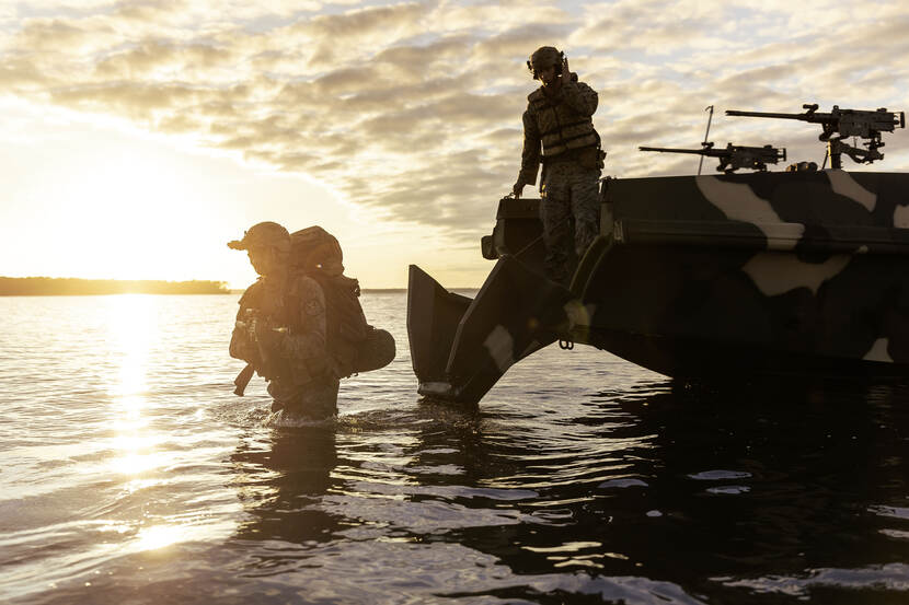 Een marinier loopt door het water, dat zijn bovenbeen reikt. Achter hem de boot, waar hij zojuist is afgestapt. Op de achtergrond is de ondergaande zon te zien.