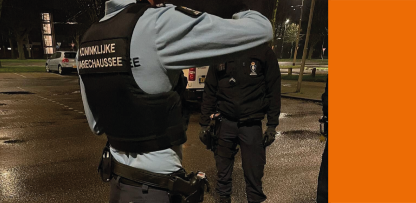 2 leden van de KMar zijn op straat met elkaar in gesprek. Met op de achtergrond bomen en 2 auto’s