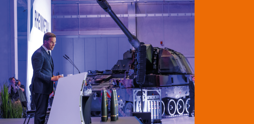 SG NAVO Mark Rutte achter een spreekgestoelte bij Rheinmetall, met een pantserwagen op de achtergrond.