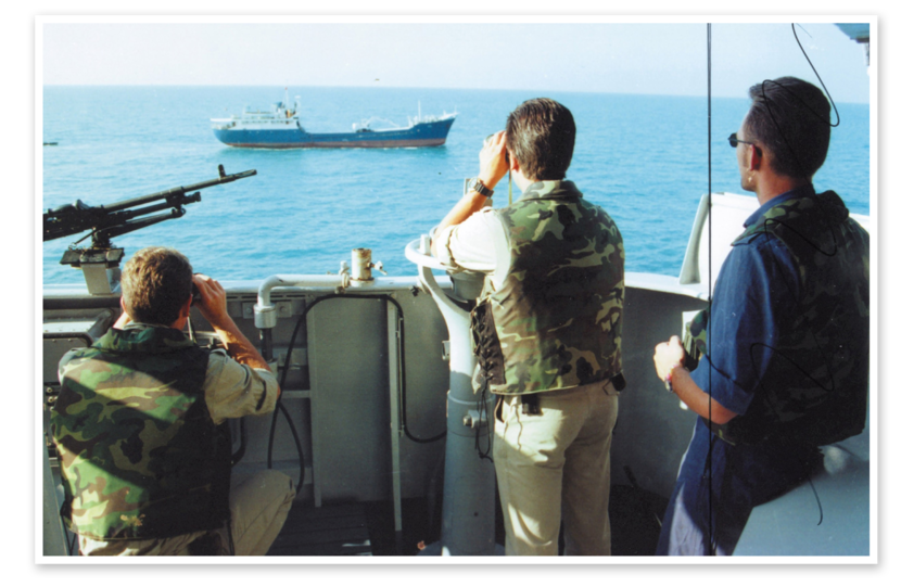 Drie personen staan naast elkaar bij de rand van een schip. Twee van hen kijken door verrekijkers naar een koopvaardijschip, dat verderop op zee vaart.