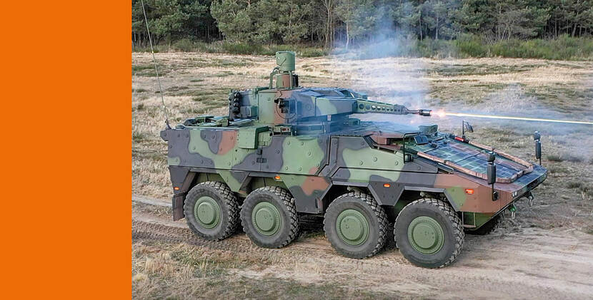 Een zwaar militair pantservoertuig in camouflagekleuren, met zes grote wielen, rijdt vurend met het kanon over een onverhard pad.