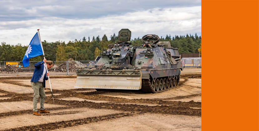 Staatssecretaris Tuinman staat op een onverhard, modderig terrein en houdt een blauwe vlag met een wit symbool vast. Hij draagt een blauw jasje en lichtgekleurde broek. Op de achtergrond staat een zwaar militair voertuig.