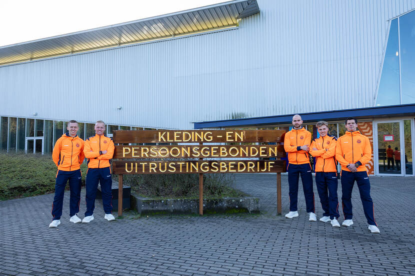 Vijf mannen in oranje sportjassen en donkerblauwe broeken staan glimlachend voor een houten bord van Kleding- en Persoonsgebonden Uitrustingsbedrijf.