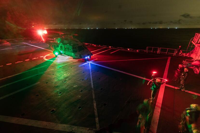Een helikopter landt op het helikopterdek van een schip. Buiten is het donker, de groene en rode lichten komen van de helikopter