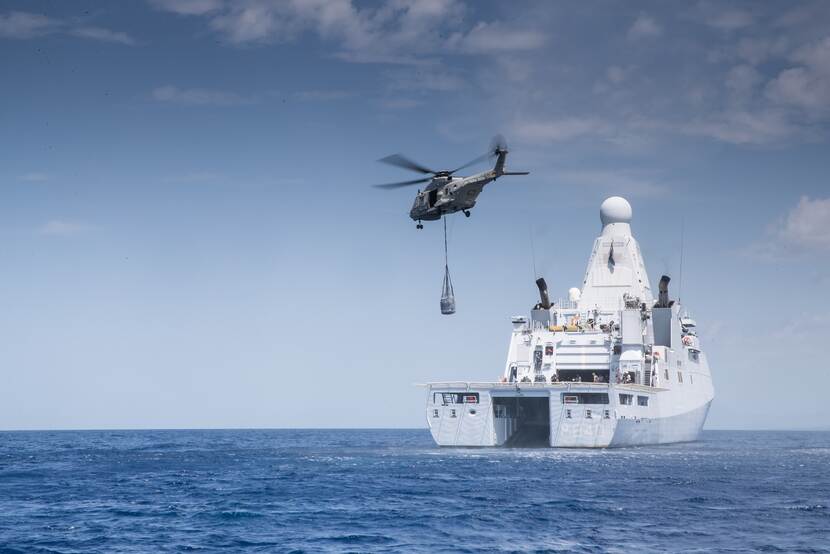 Een marineschip vaart over zee bij Haïti. Boven het dek vliegt een helikopter waaronder een box met hulpgoederen hangt.