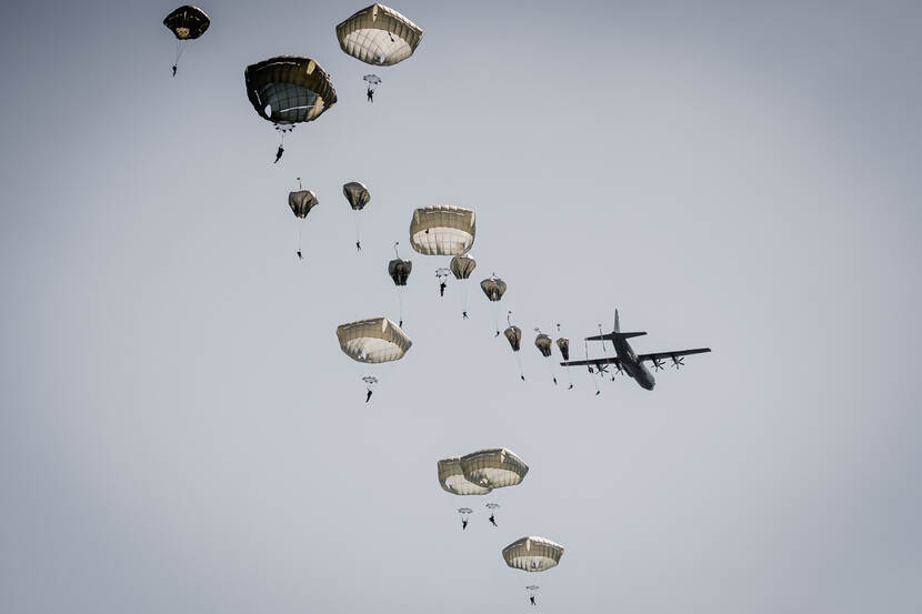 Zo’n 20 parachutisten springen uit een vliegtuig en hangen naast elkaar in de lucht.
