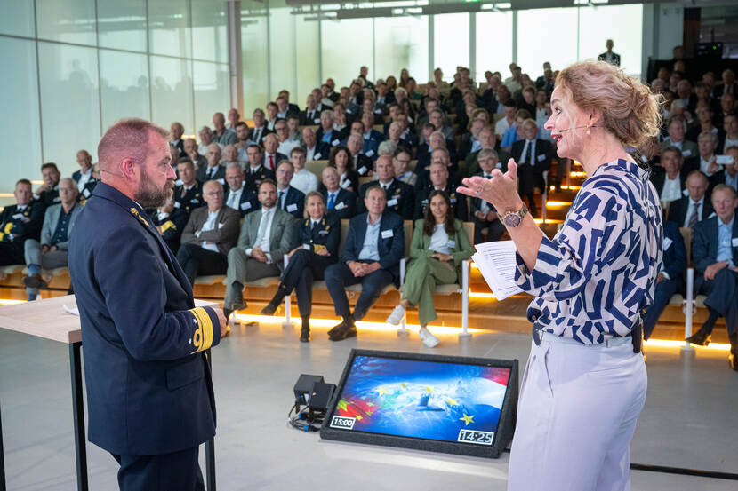 Vice-admiraal Tas praat met een dame voor een beeldscherm met op de achtergrond toeschouwers.