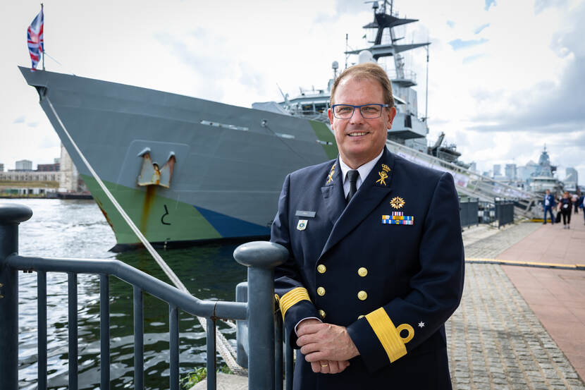 CDR Paul Flos staat met zijn handen over elkaar voor een metalen reling. Achter hem het Britse fregat HMS Iron Duke in de haven en een stadsgezicht op de achtergrond.