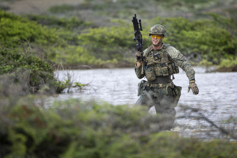 een militair met zijn persoonlijk wapen omhoog gericht rennend door een plas water.