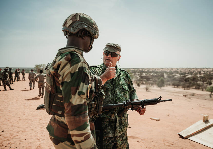 Een Nederlandse militair met zonnebril legt met handgebaren iets uit aan een Afrikaanse militair.