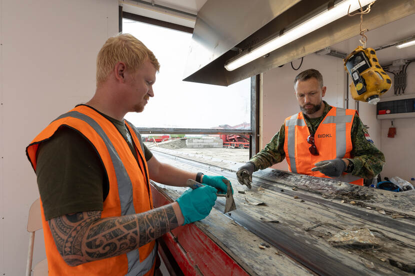 De lopende band in de container, van binnen gezien. Twee militairen buigen over het voorbijglijdende materiaal, met een schepje in de hand. Beide militairen dragen een oranje veiligheidsvest en handschoenen.