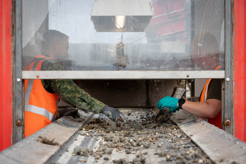 Een lopende band, met achter een plexiglas luik twee militairen die voorbijkomend materiaal selecteren. De een heeft een oranje vest aan en wijst naar iets dat voorbij komt.