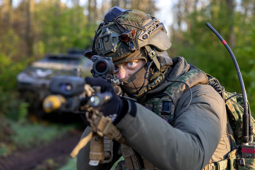 Een militair kijkt dreigend langs de camera, met een lang wapen getrokken voor zijn gezicht. Achter hem staat een Fennek-verkenningsvoertuig.