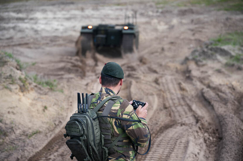 Een onbemenst rijdend rupsvoertuig rijdt door een zanderig oefenterrein. Op de voorgrond een militair, vanaf de rug bezien, die het voertuig bestuurt.