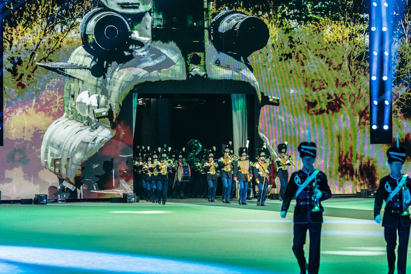 De Regimentsfanfare Garde Grenadiers en Jagers betreedt de vloer alsof ze uit een hoverende Chinook stappen. De helikopter is met openstaande laadklep op het LED-scherm geprojecteerd.