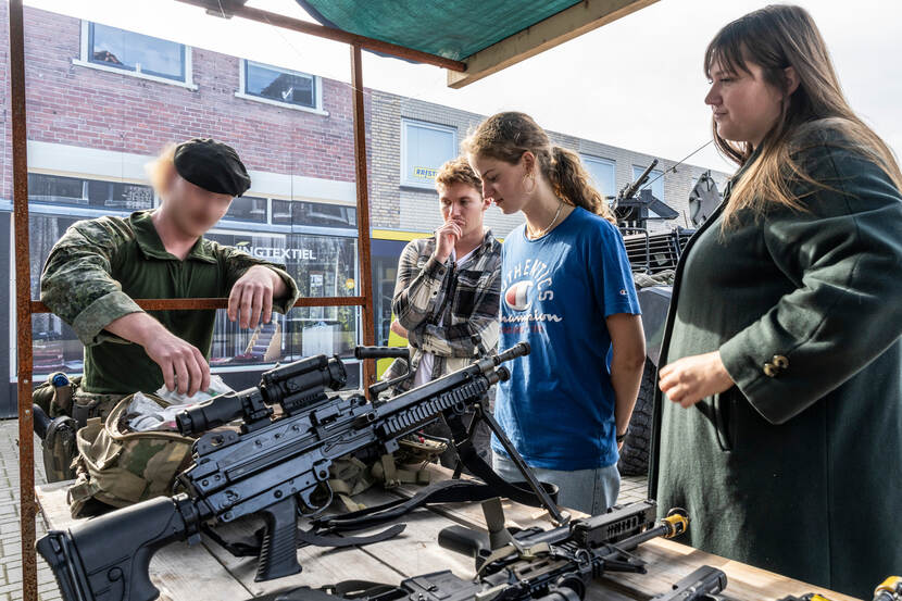 Een militair buigt over een kraampje, waarop verschillende wapens te zien zijn, twee toeschouwers kijken mee.