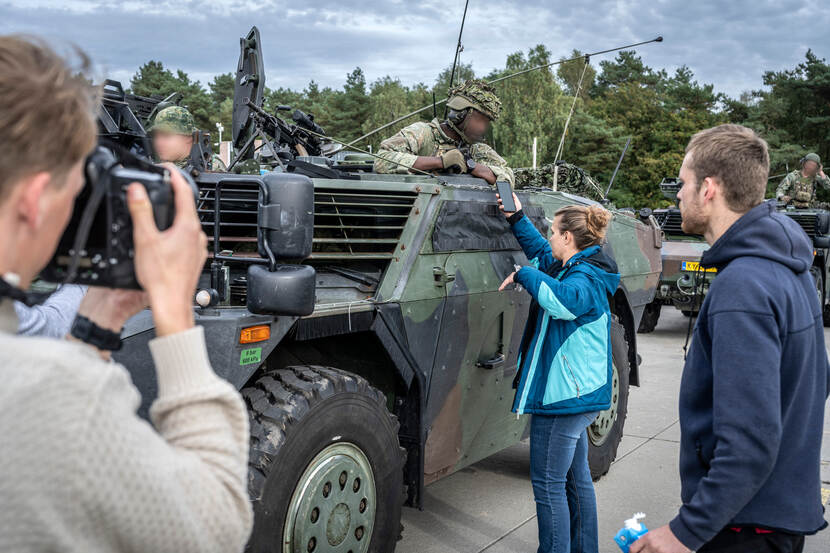 Twee militairen hangen uit de Fennek. Om het verkenningsvoertuig drommen de mensen, waarvan een met een grote fotocamera in de hand, de ander neemt quotes op met de telefoon.