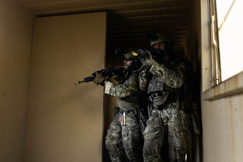 Twee Militairen bewegen met getrokken lange wapens door gebouw in het oefendorp.