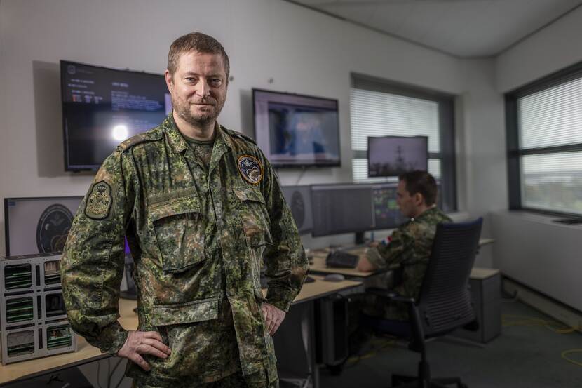 Militair in uniform staat voor een aantal schermen. Naast hem zit een collega aan een bureau.