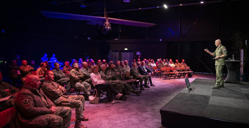 Publiek luistert naar een militair spreker op een podium.