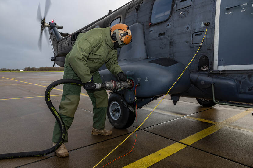 Een militair tankt een helikopter met draaiende motor en rotorbladen bij op een helikopterplatform.