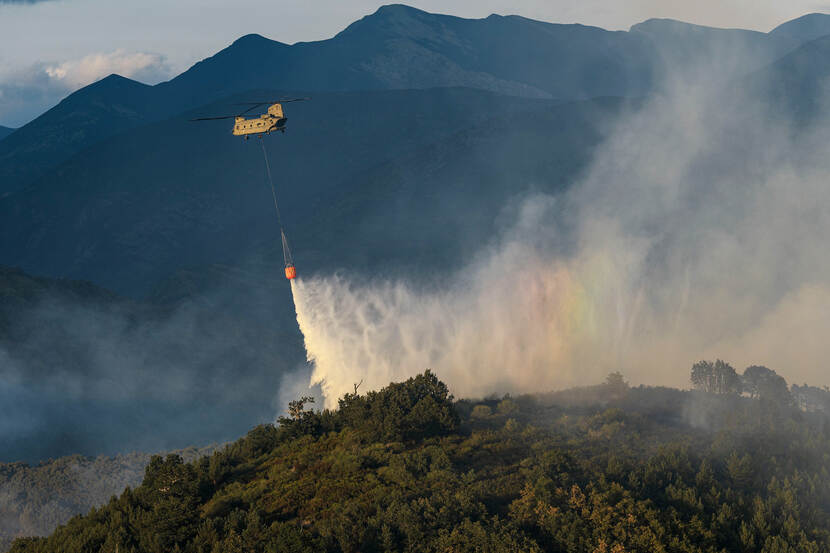 Een Chinook, met daaronder een Bambi Bucket, gooit water over een bosbrand.