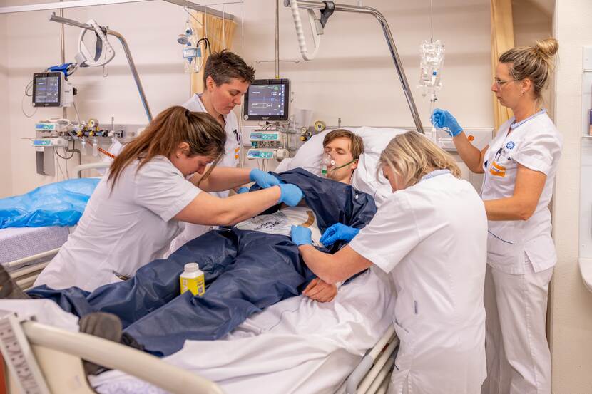 Vier verpleegkundigen in witte kleding rondom een patiënt in een ziekenhuisbed.