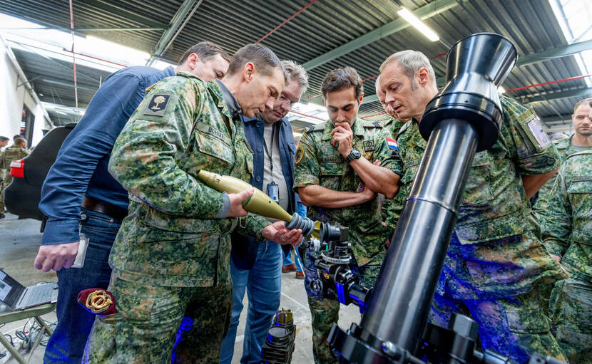 Mensen in burger en militairen kijken en luisteren naar de uitleg bij een mortier.