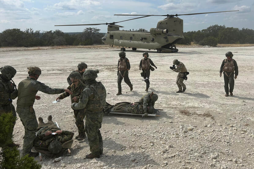 Militairen op onverhard buitenterrein dragen twee militairen liggend op brancards over aan collega-militairen met op de achtergrond een Chinook.