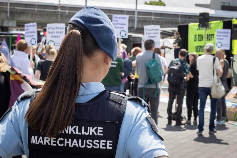 Een vrouwelijke Marechausseecollega op de rug gezien, op de achtergrond lopen demonstranten.