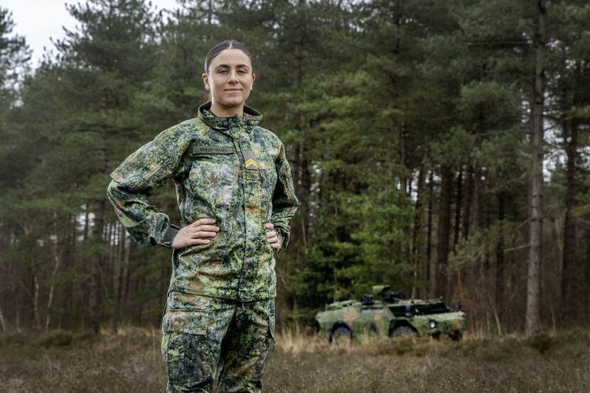 Een vrouwelijke militair in het nieuwe gevechtspak met op de achtergrond een verkenningsvoertuig en bomen.