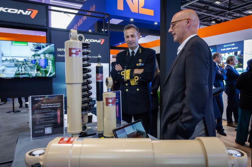 Vice-admiraal Jan Willem Hartman staat met zijn armen over elkaar en heeft een kleine glimlach. Ondertussen is hij in gesprek met een andere man in pak. De twee kijken naar een beige-kleurig object op een tafel.