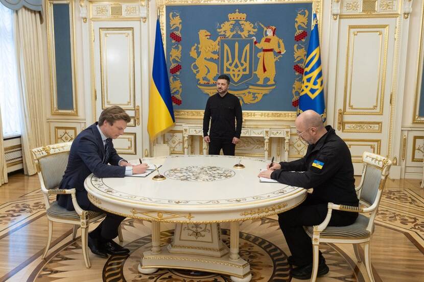 Defensieminister Ruben Brekelmans en zijn Oekraïense collega Shmyhal zitten aan een ronde tafel en zetten beiden een handtekening. Daarachter Oekraïense vlaggen met daartussen een toekijkende president Zelensky.