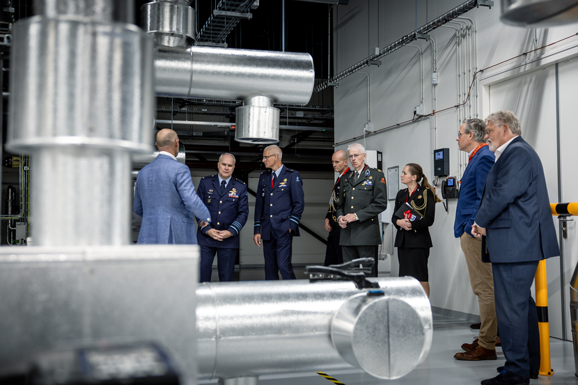 Generaals Eichelsheim en Boekholt luisteren naar een man in pak. Daarnaast staan meerdere personen die hem ook aanhoren. Op de voorgrond zijn zilverkleurige buien te zien.