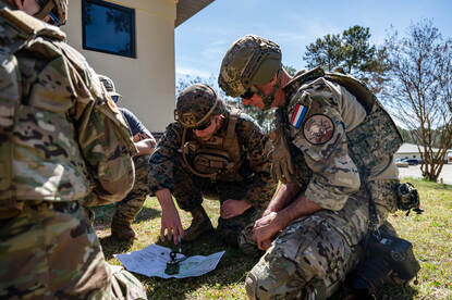Drie militairen zitten gehurkt in het gras en bekijken een landkaart.