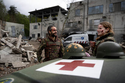 Twee oefenslachtoffers met nagemaakte wonden staan achter een militaire  ziekenwagen, voor het oefengebouw.
