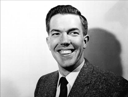 Zwart-wit portret van een man die langs de lens kijkt met een brede lach waarbij veel tanden zichtbaar zijn. Haar strak naar achteren, jasje en dasje.