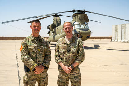 Sergeant-majoor Dennis en korporaal Tim kijken in de camera op Al Asad Airbase in Irak. Op de achtergrond staat een geparkeerde Chinook-transporthelikopter.