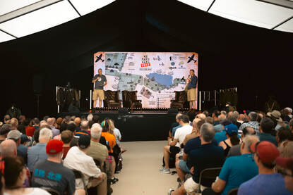 Mensen luisteren aandachtig naar twee mannen op het podium, met tussen hen in een grote map van het festivalterrein.