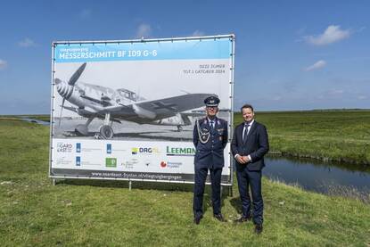 Kolonel Volker Eden en de Friese burgervader Johannes Kramer poseren voor de camera voor een groot bord, waarop een oude foto van een Messerschmitt (het neergestorte vliegtuig) staat afgebeeld in zwart-wit.