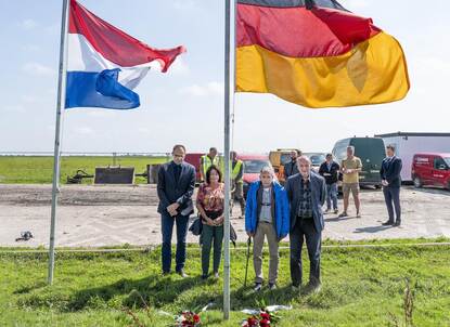 Een wapperende Nederlandse en Duitse vlag in de kwelder nabij het Friese Hallum, met daarachter mensen die een minuut stilte houden.