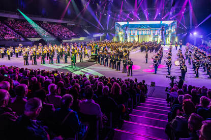 Voornamelijk paars uitgelichte zaal waarop grote delen van de tribune zichtbaar zijn met in het midden de volledige vloer waarop alle taptoedeelnemers staan tijdens de finale.