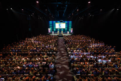 De theaterzaal van evenementenlocatie Hart van Holland, van bovenaf bezien. De zaal is gevuld met onderofficieren, die op stoelen zitten. Op het podium staan sprekers in een gevechtspak.