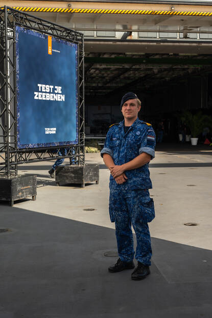 Een marineman poseert met handen over elkaar voor een promotiedoek van de marine.
