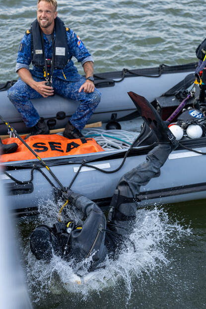 Een duiker gaat achterover het water in, terwijl een collega in de rubber boot toekijkt.
