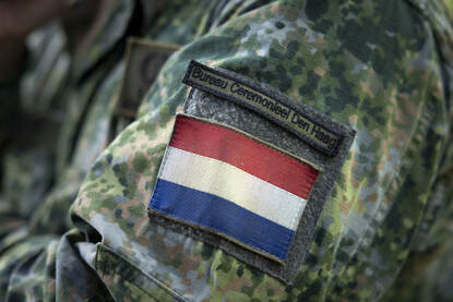 Een mouw van een militair pak met daarop een Nederlandse vlag en daarboven een ‘straatnaambord’ met de tekst ‘Bureau Ceremonieel Den Haag’.