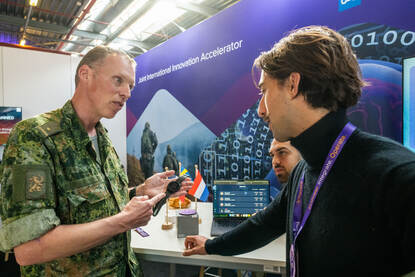 Links een horloge met veel letters, rechts een militair die spreekt met twee mannen aan een tafel met de vlaggetjes van Nederland, Frankrijk en Zweden.