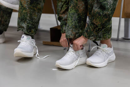 Twee personen dragen witte, moderne sportschoenen met een dikke, gebogen zool en een donkerblauwe broek met oranje strepen langs de zijkant. Rechts: Twee personen in camouflagebroek dragen witte, moderne sportschoenen met dikke zolen; een persoon bindt de veters vast,.