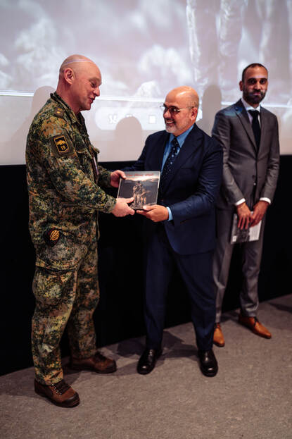 Man in pak overhandigt boek aan militair. Een tweede man in pak kijkt toe.